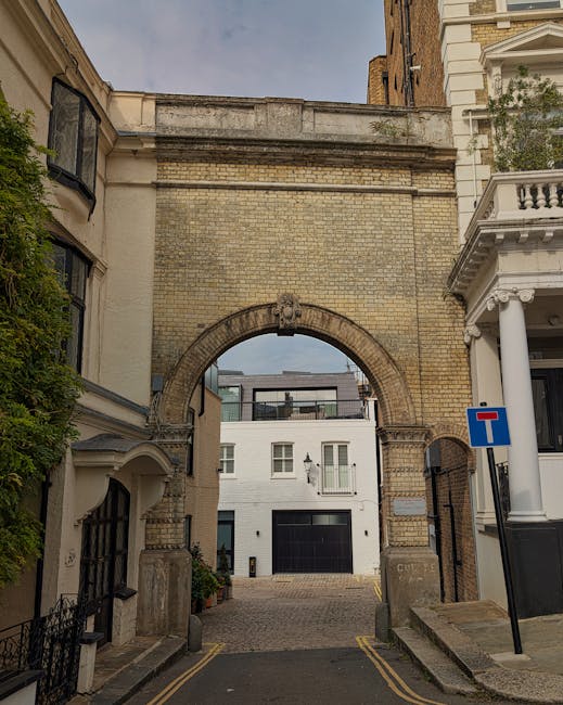 A narrow urban street in Kensington, with a large brick archway overhead connecting two historic buildings, one featuring classical white columns and decorative balustrades. The street is paved with c