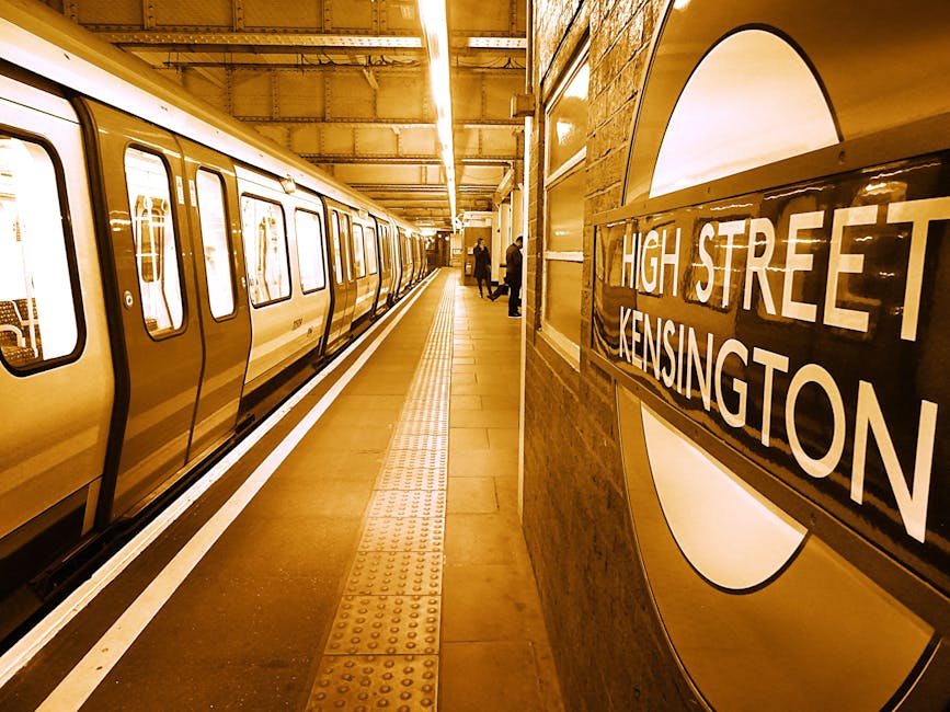 A narrow urban street in Kensington, with a large brick archway overhead connecting two historic buildings, one featuring classical white columns and decorative balustrades. The street is paved with c