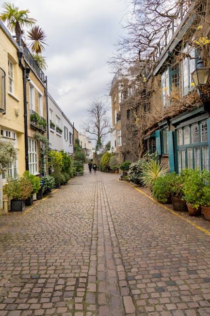 A narrow residential street in South Kensington SW7 lined with parked cars on both sides, including small hatchbacks, sedans, and vans, with pink flowering trees with blossoms on the branches overhang
